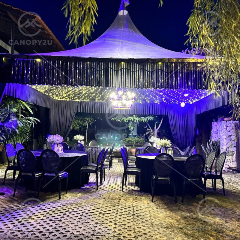 Outdoor nighttime event under a white tent with twinkling string lights, round tables, black chairs, and a glowing chandelier among plants.